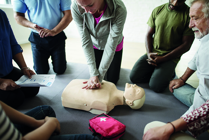 Sesión de entrenamiento de reanimación cardiopulmonar (RCP) en grupo. Un grupo de personas sentadas en círculo realiza un entrenamiento de RCP en un maniquí, con una instructora practicando compresiones torácicas y un kit de primeros auxilios visible.