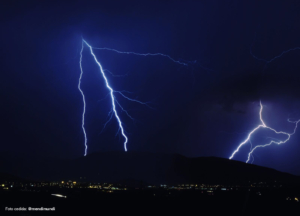 Fotografía nocturna de la Vía Láctea con una descarga eléctrica (rayo) en el horizonte sobre un paisaje de montaña.